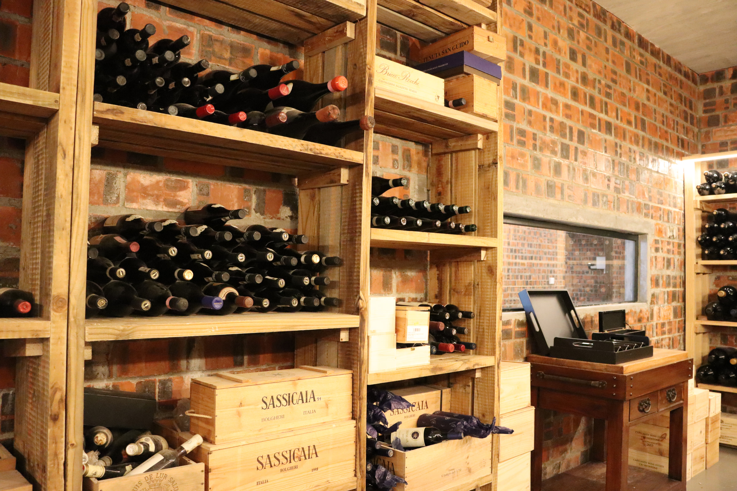 Wooden racks of wine bottles in the cellar, with Sassicaia cases stacked below against a brick wall