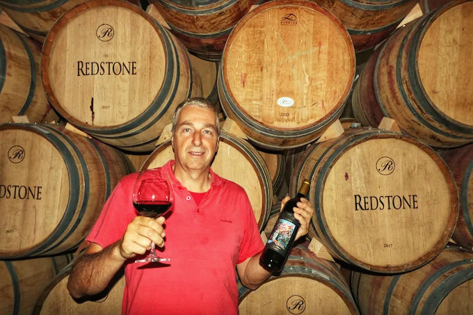 Johann Innerhofer raising a glass and a bottle of Redstone in front of barrels stencilled Redstone