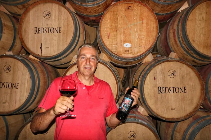 Johann Innerhofer raising a glass and a bottle of Redstone in front of barrels stencilled Redstone